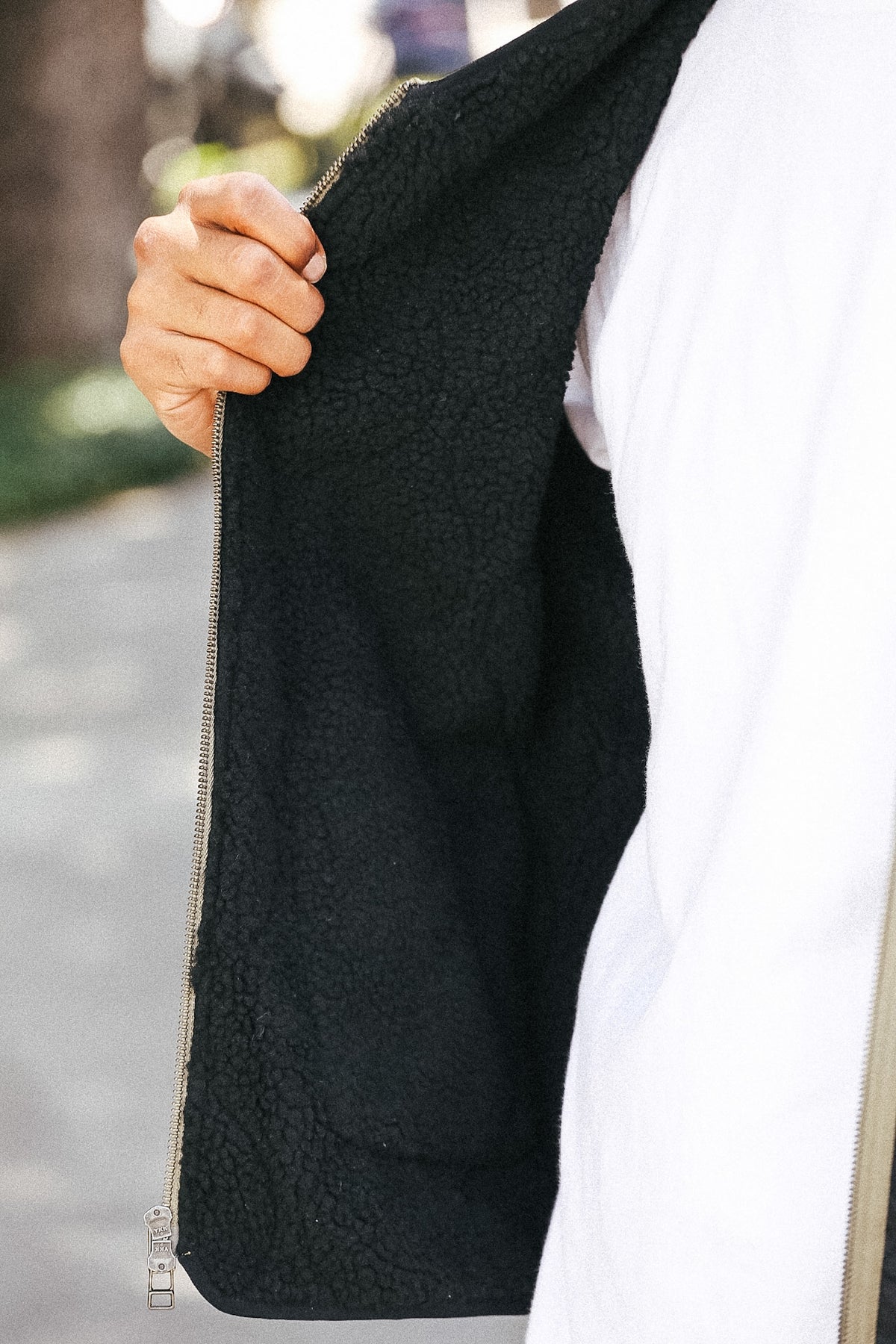 Close-up of a person wearing a black sherpa-lined zip jacket over a white shirt, holding the zipper open with a hand outdoors.