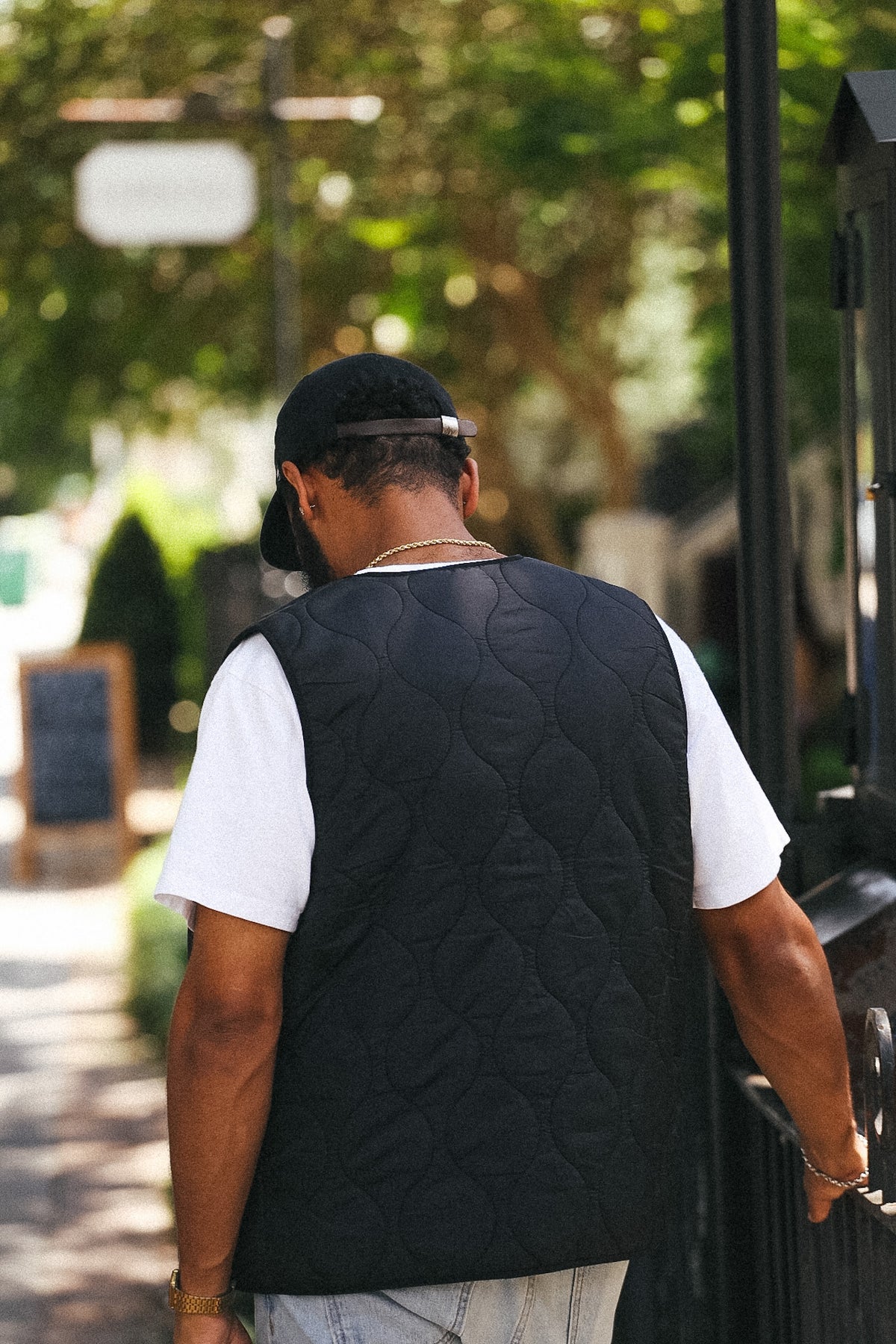 A man viewed from behind wearing a black quilted vest over a white t-shirt and a black baseball cap, walking outdoors on a sunny day.
