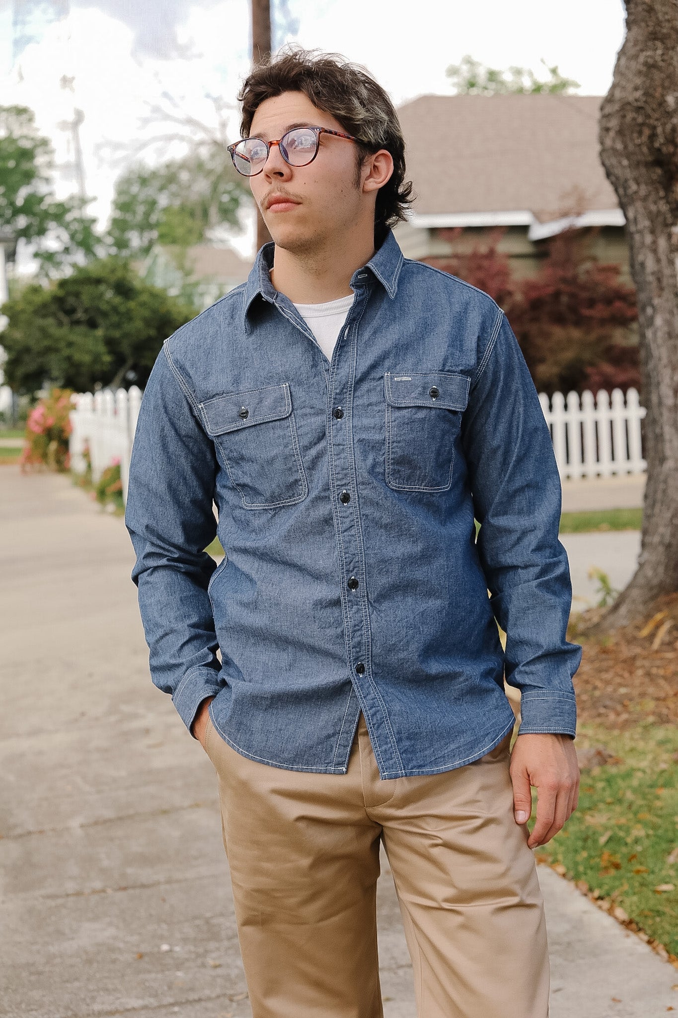 Man wearing a blue denim shirt and beige pants standing on a sidewalk.