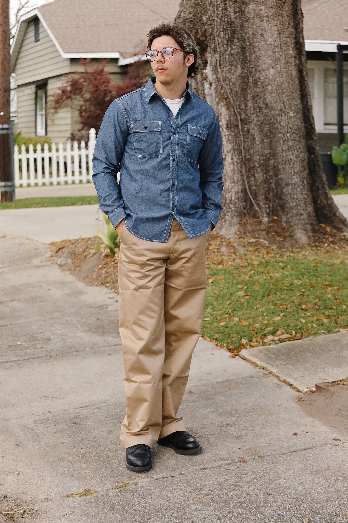 Man wearing a blue denim shirt and beige pants standing on a sidewalk with a tree and house in the background.