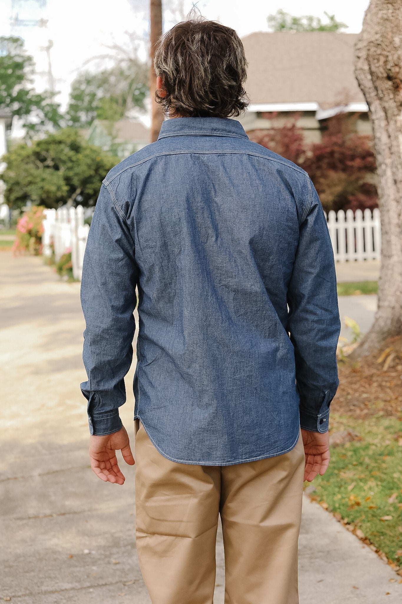 Man wearing a blue denim shirt and beige pants walking on a sidewalk.
