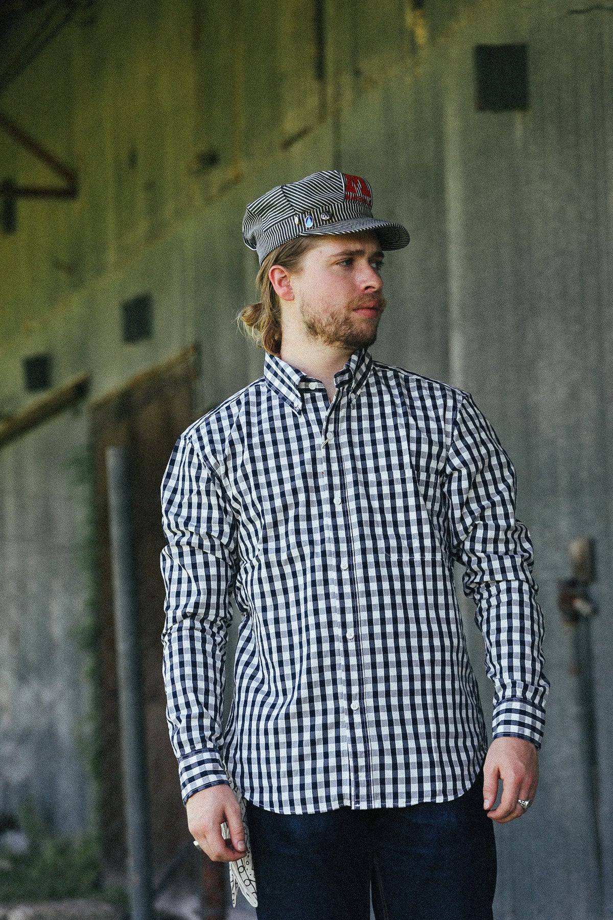 A man wearing a black and white gingham long-sleeve button-down shirt and a matching gingham cap with a red logo patch, standing in front of a rustic concrete wall.