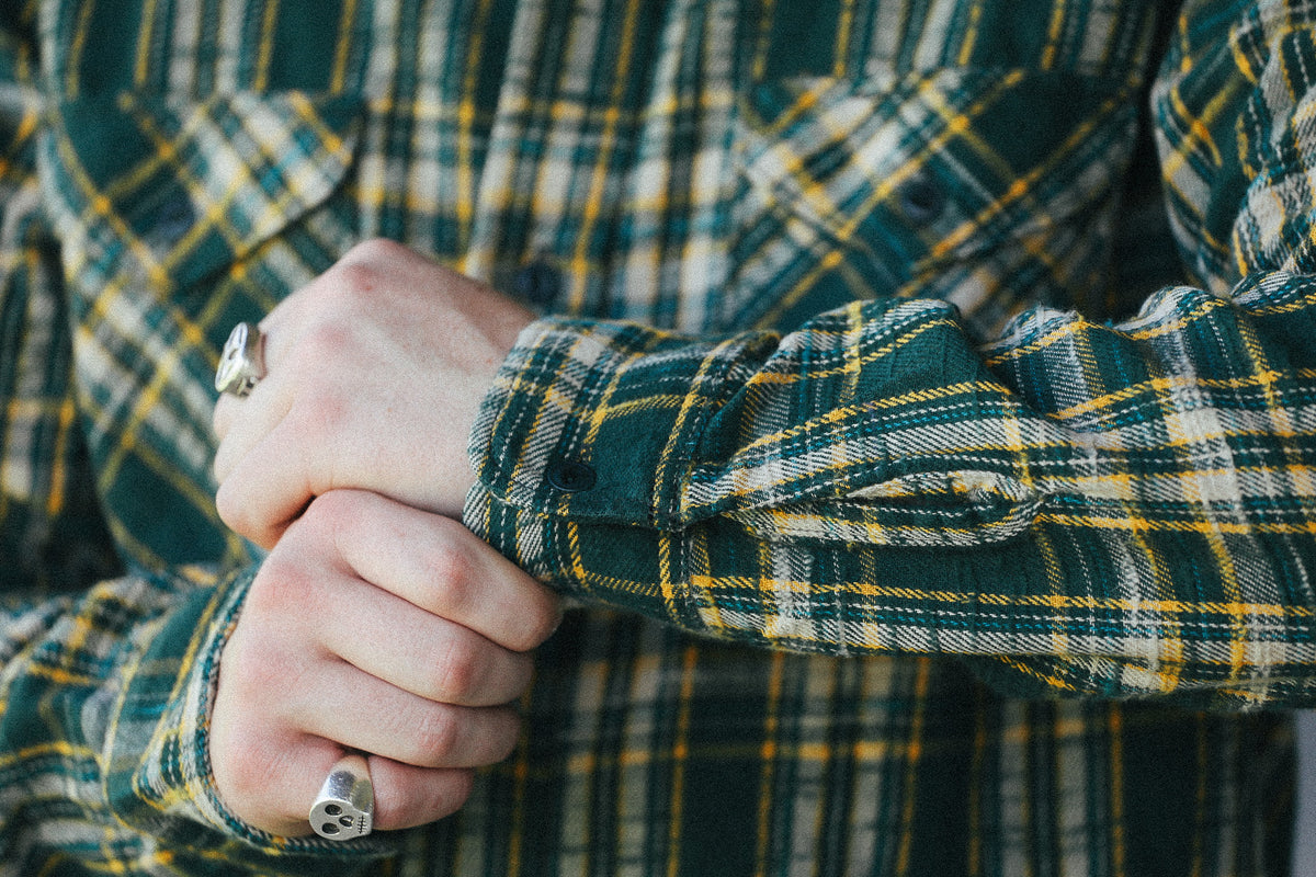 A close-up of a person's hands clasped in front of a green and yellow plaid flannel shirt. The person is wearing silver rings on both fingers.