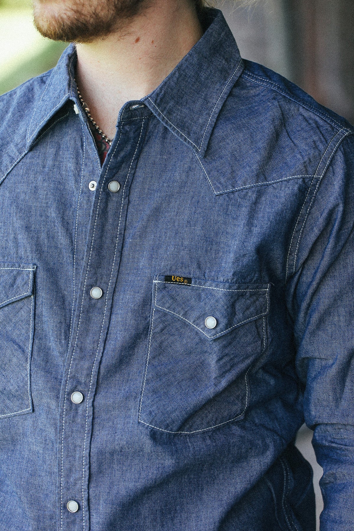 A close-up of a man wearing a blue denim western-style button-up shirt with a chest pocket and a small yellow brand tab.