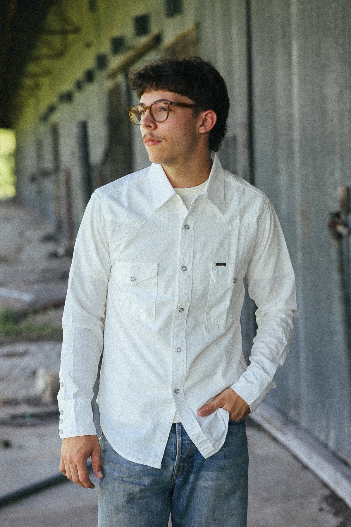 A man wearing a white long-sleeve button-up shirt with two chest pockets and blue jeans, standing in an outdoor industrial setting.