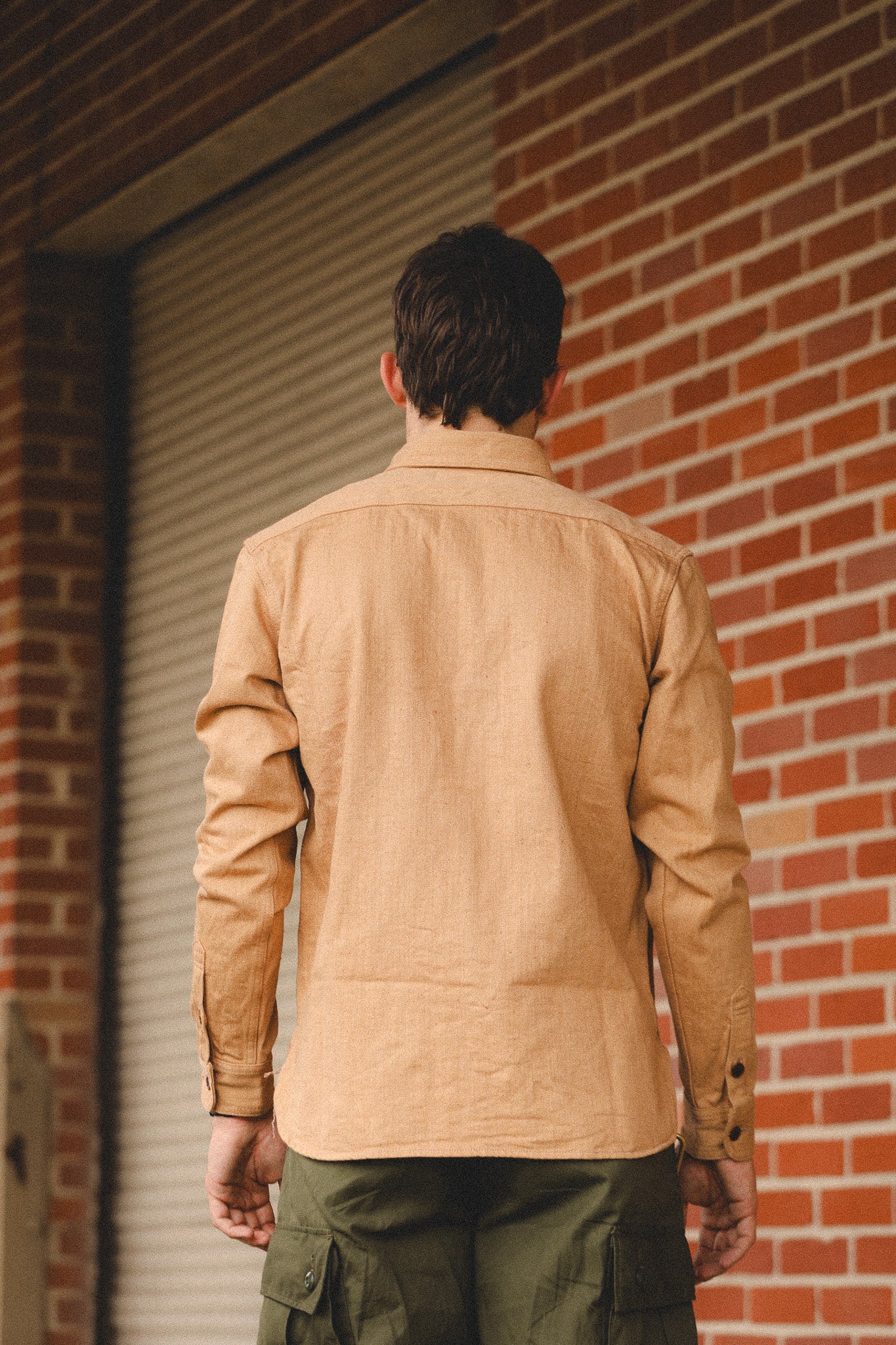 Person wearing a beige jacket and green pants standing against a brick wall.