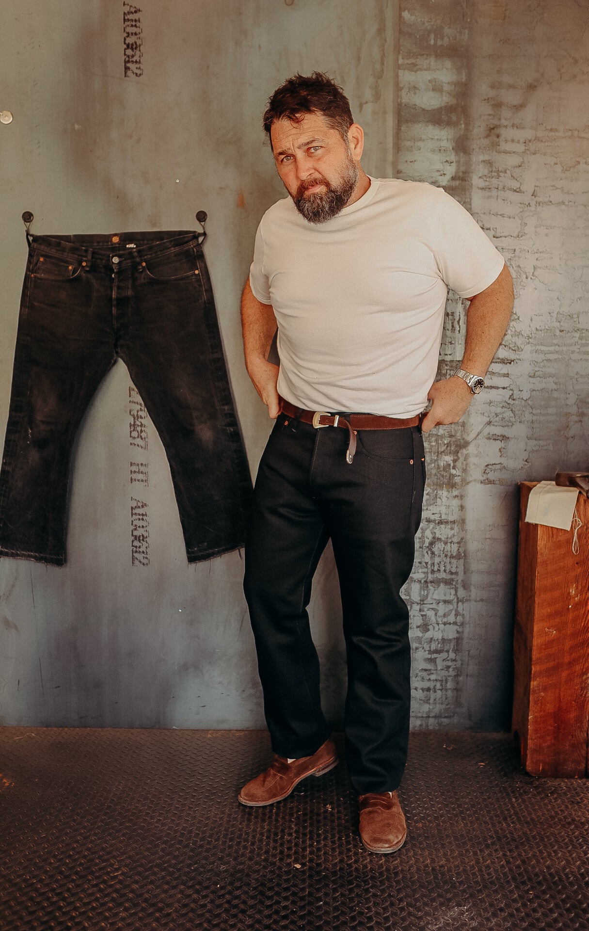 Man with beard wearing white t-shirt and black jeans standing indoors near hanging black denim jeans