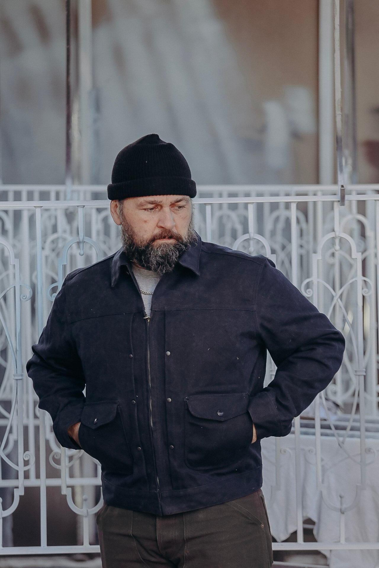 Bearded man wearing black beanie and dark jacket with pockets, standing in front of metal gate outdoors
