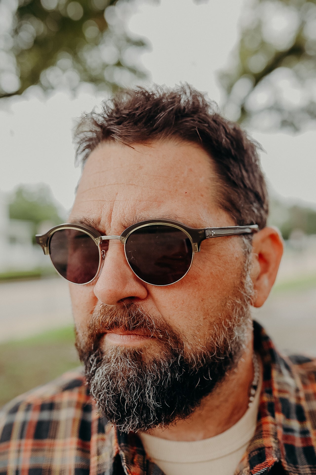 Bearded man wearing round sunglasses and a plaid shirt outdoors with blurred trees