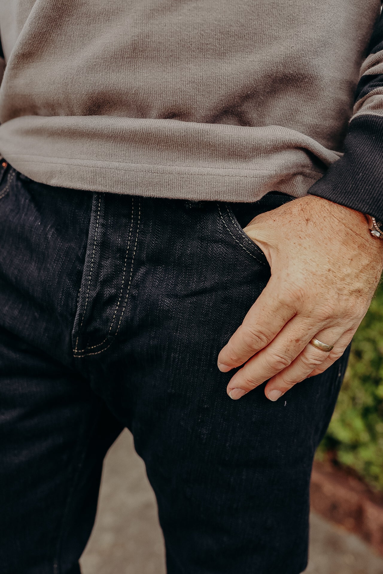 Close-up of a person wearing dark denim jeans and a beige sweatshirt with one hand in pocket and wedding ring