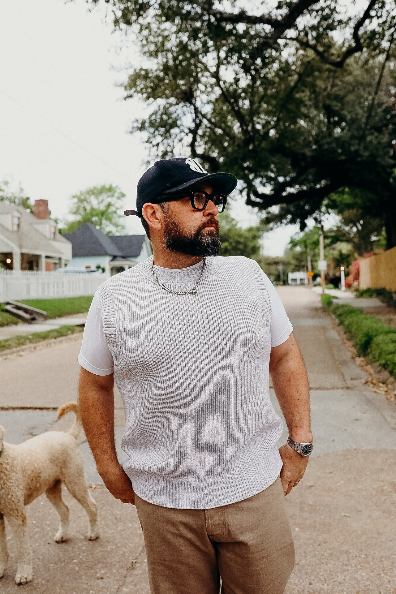 Man wearing beige knit vest, white t-shirt, black cap, and glasses standing on suburban street with dog