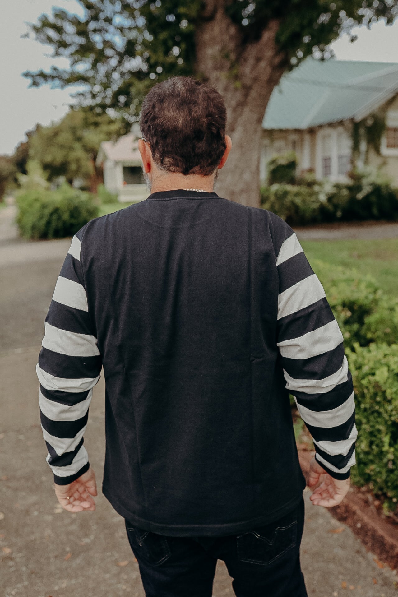 Man wearing black long-sleeve shirt with white stripes on sleeves, standing outdoors near bushes and a house