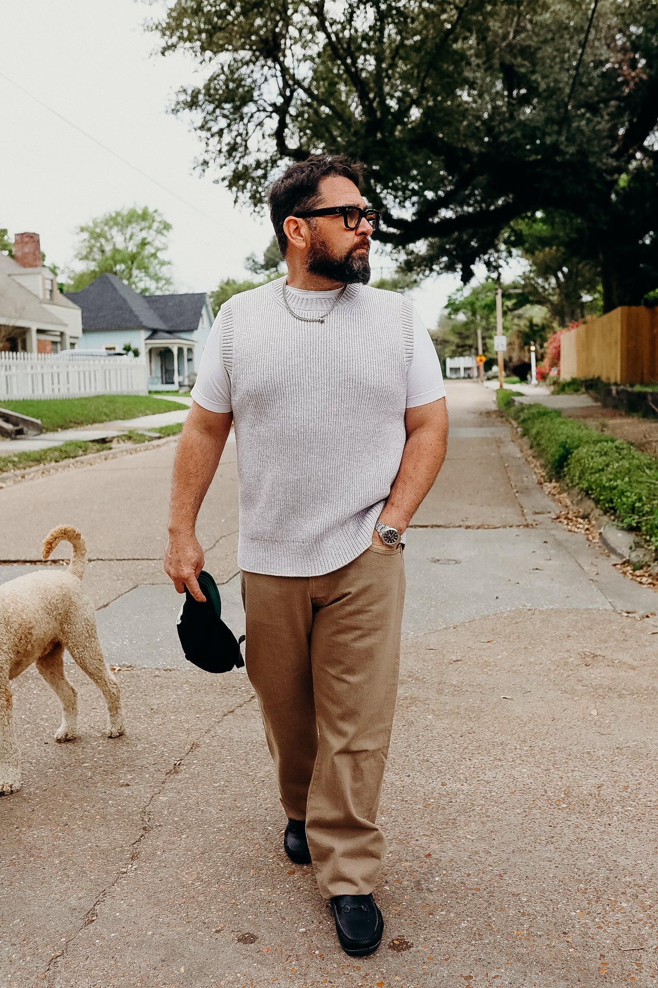 Man wearing white knit vest, beige pants, and black shoes walking a dog on a suburban street