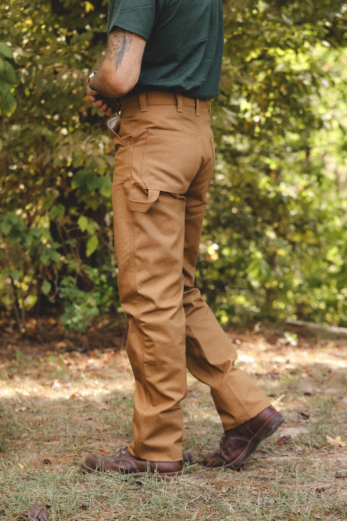 A person outdoors wearing tan cargo-style pants, a dark green shirt, and brown boots, shown from the back in a wooded setting.