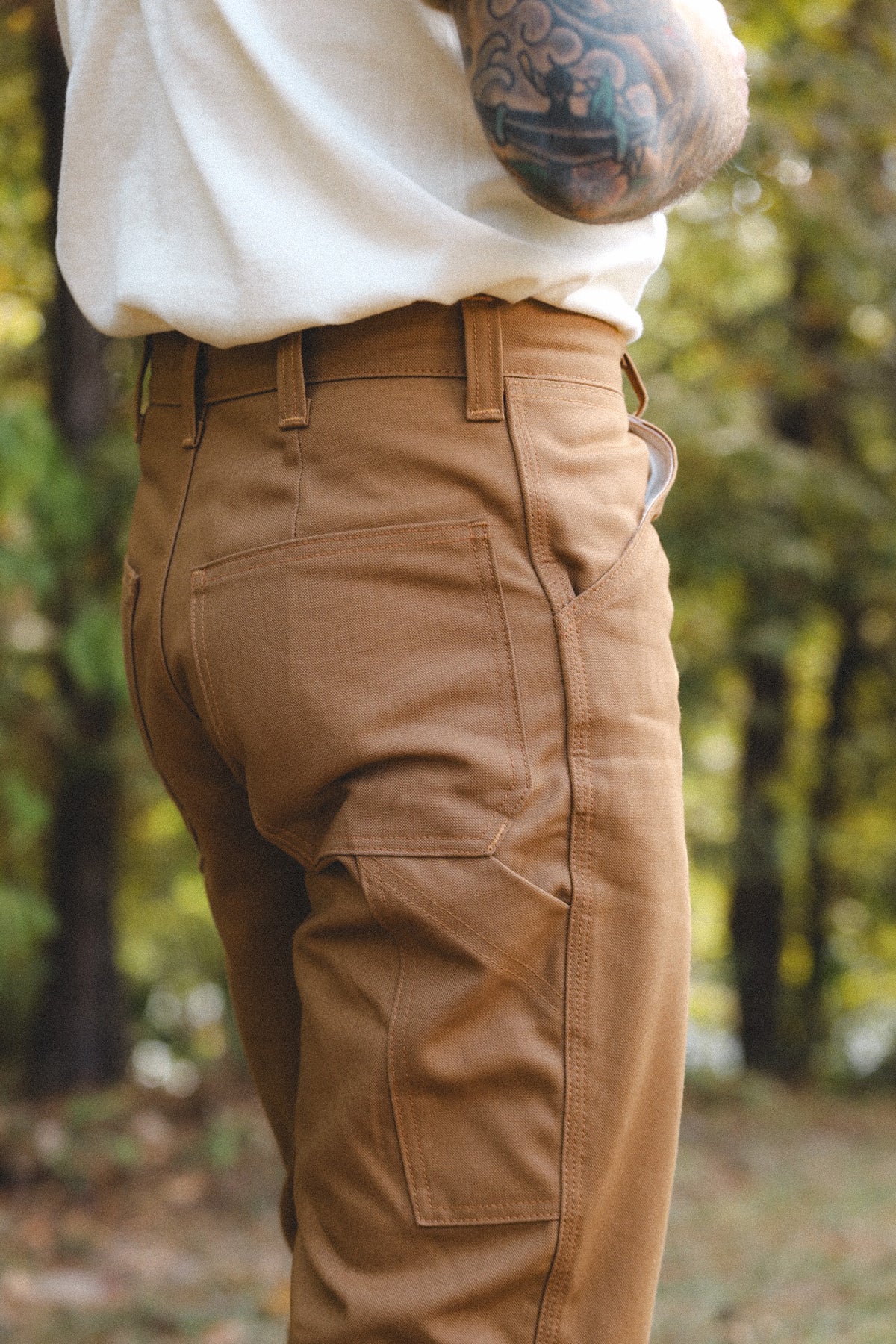 A close-up side view of a person wearing tan cargo pants and a white shirt, standing outdoors in a blurred forest setting.