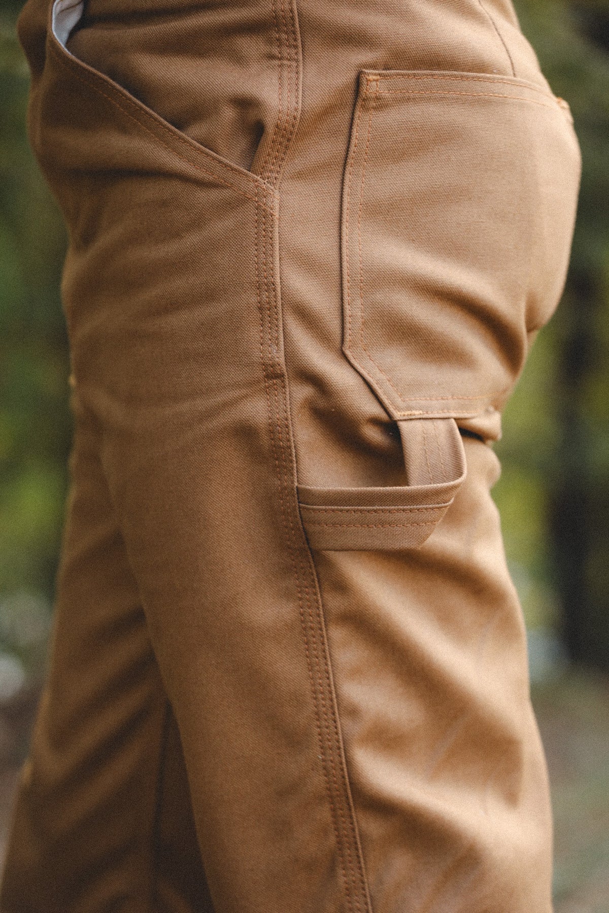 Close-up of tan work pants showing the side pocket and cargo-style flap pocket, worn outdoors with a blurred natural background.
