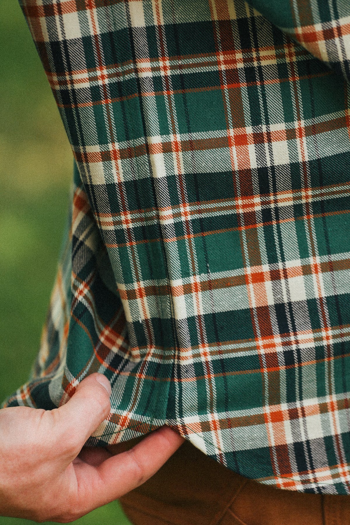 Close-up of a person wearing a green, white, and orange plaid flannel shirt, with a hand pulling the bottom hem slightly to show the fabric texture.