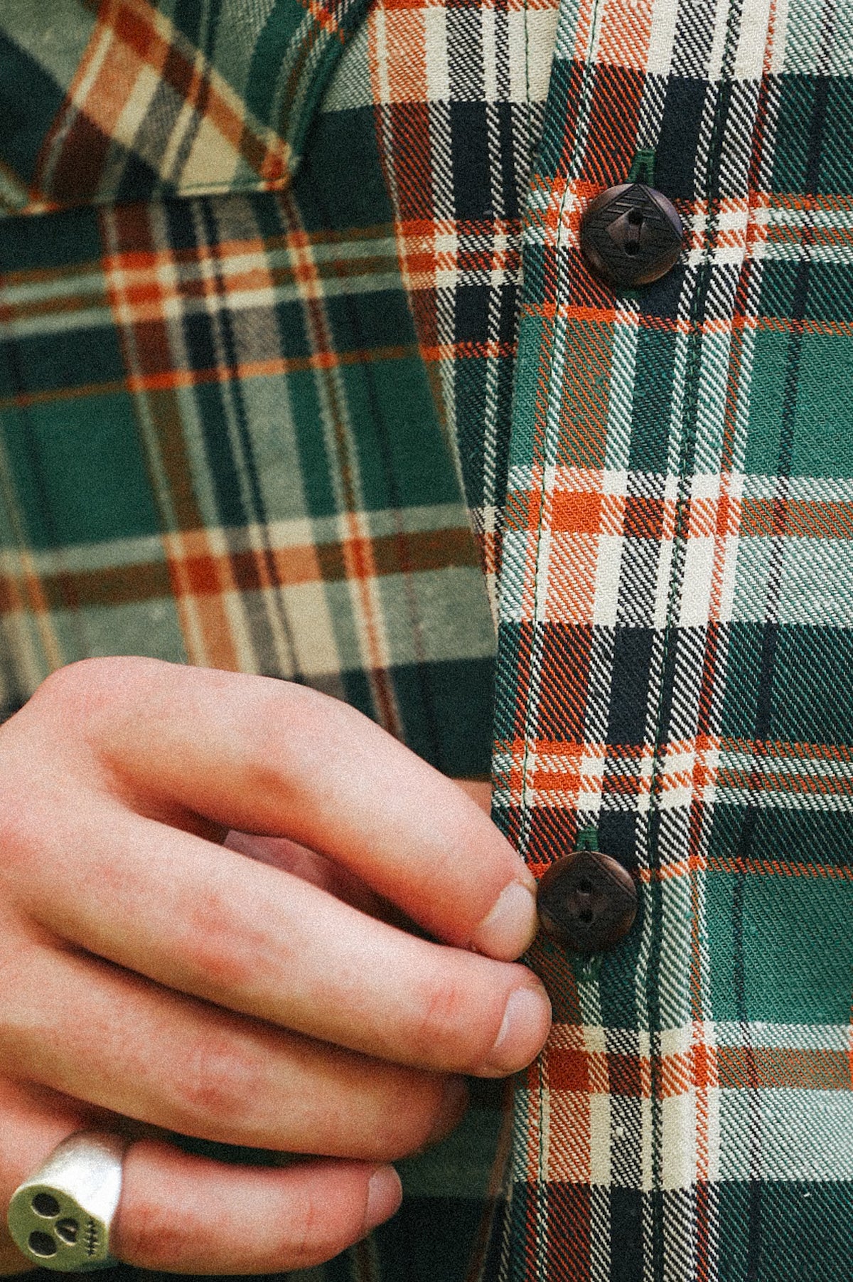 Close-up of a person's hand fastening a dark brown button on a green, orange, and white plaid flannel shirt.