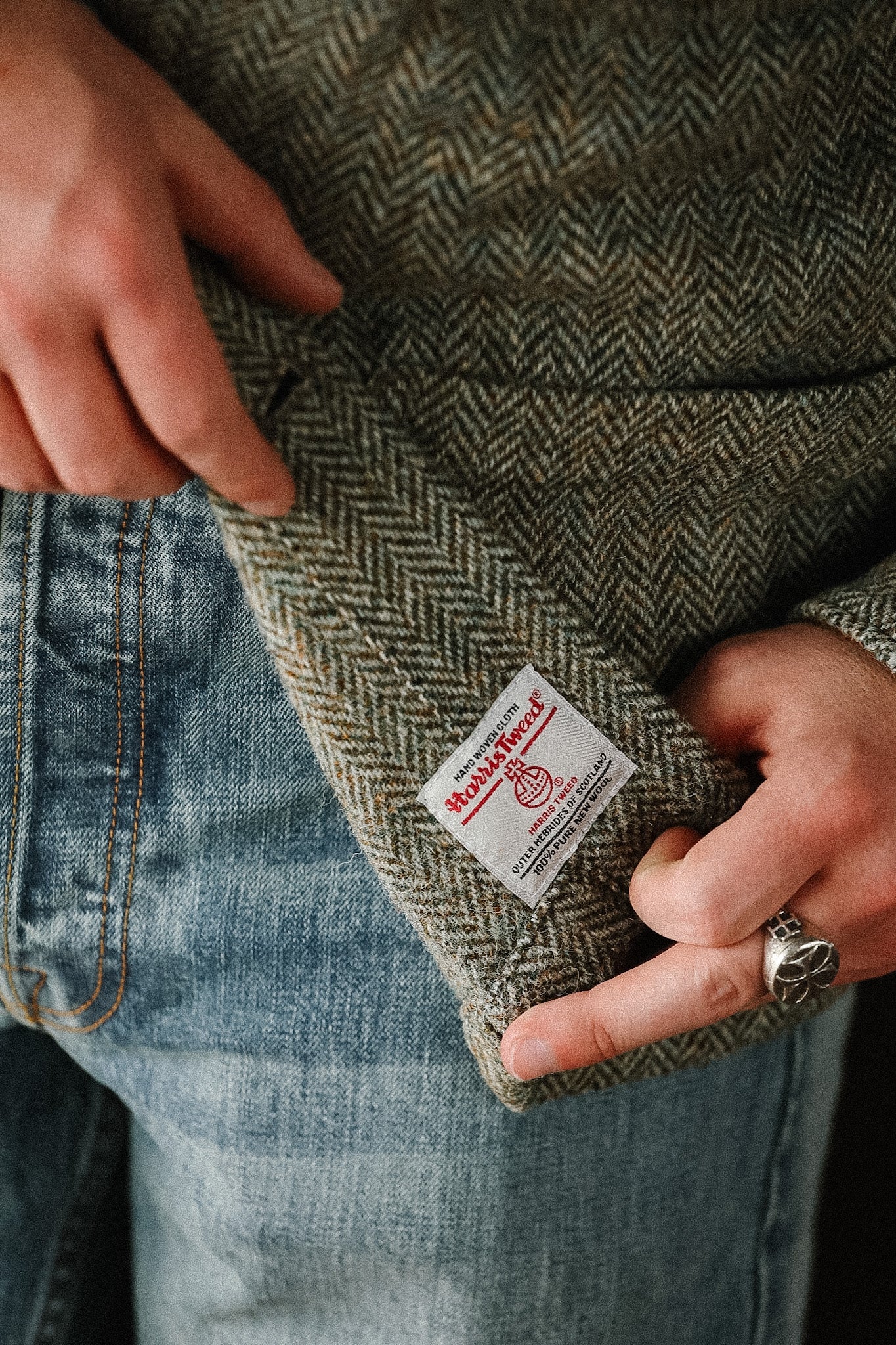 Close-up of a person wearing a green herringbone jacket with a brand label, holding the fabric.