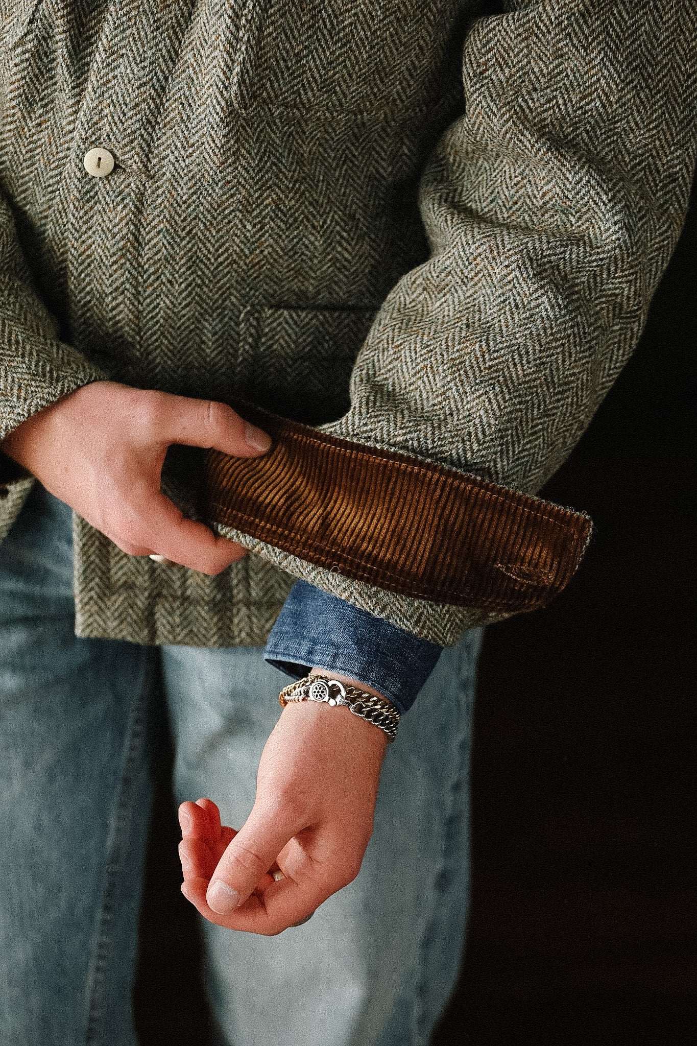 Person wearing a green herringbone jacket with a brown leather patch on a dark background