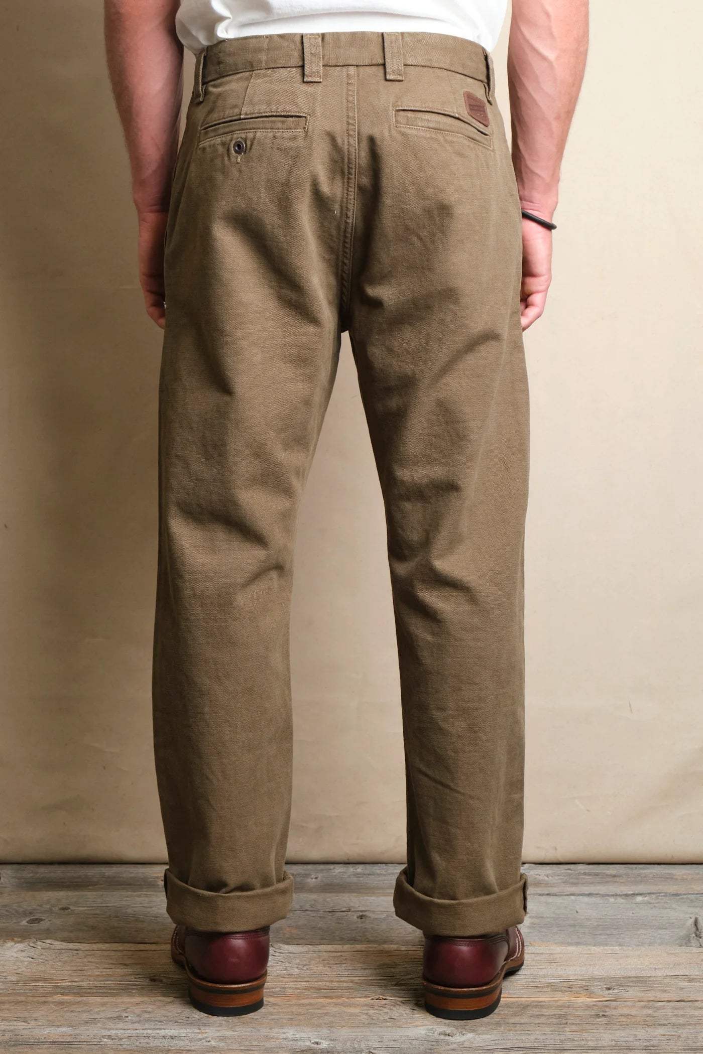 Man wearing rolled-up olive green casual pants and brown leather boots standing on wooden floor
