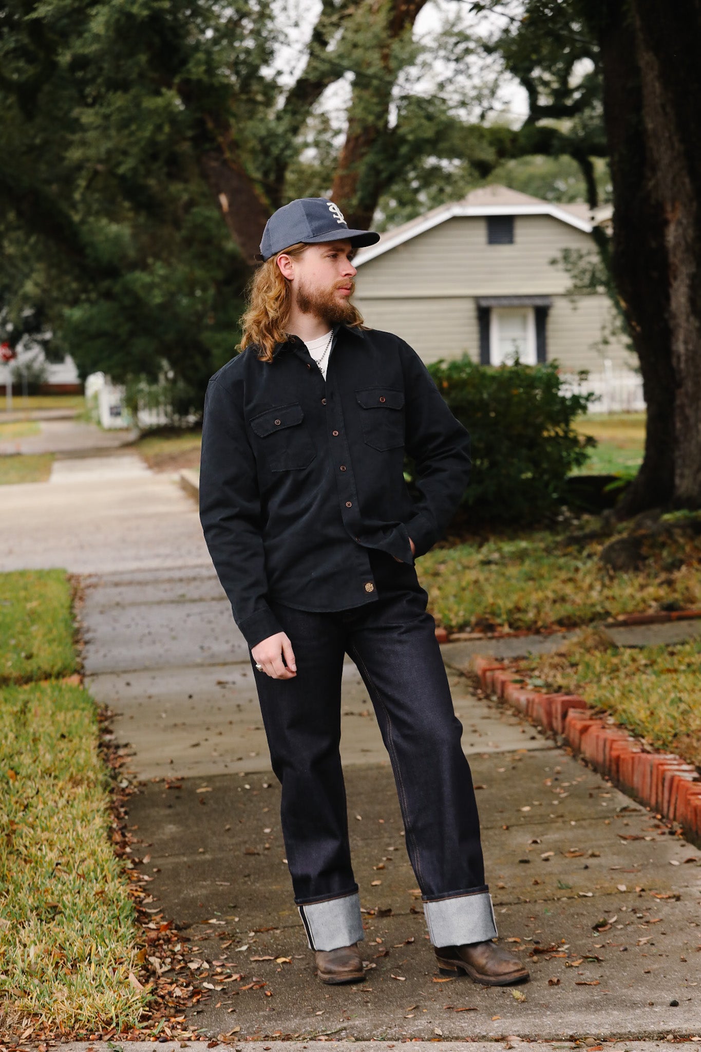 Man with long hair and beard wearing a dark jacket and jeans standing on a sidewalk.