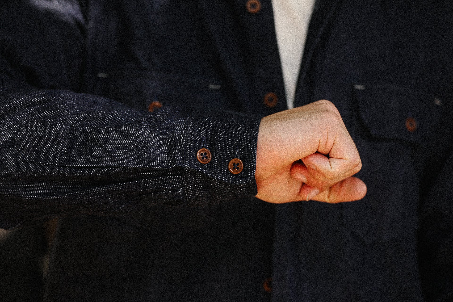Close-up of a person's fist wearing a dark denim shirt with brown buttons from Iron Shop Provisions