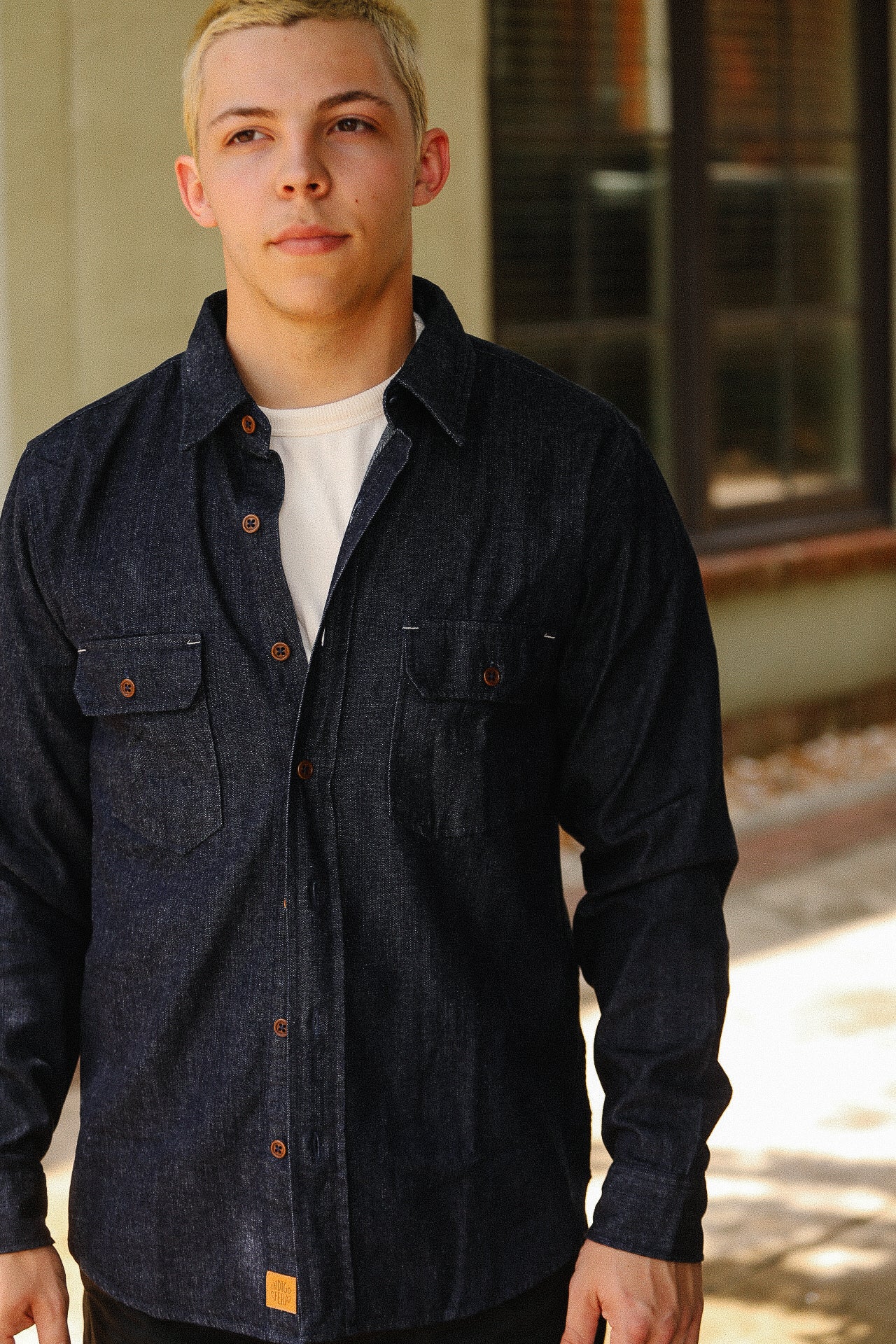 Man wearing dark denim shirt with brown buttons over white t-shirt standing outdoors near building with window