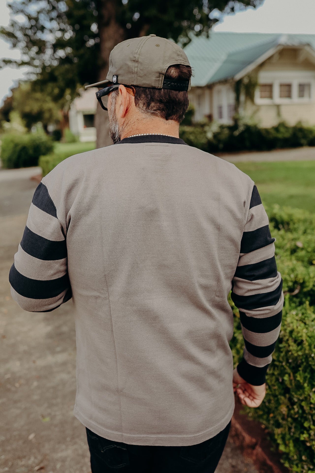 Man wearing gray long-sleeve shirt with black stripes and green cap outdoors near house