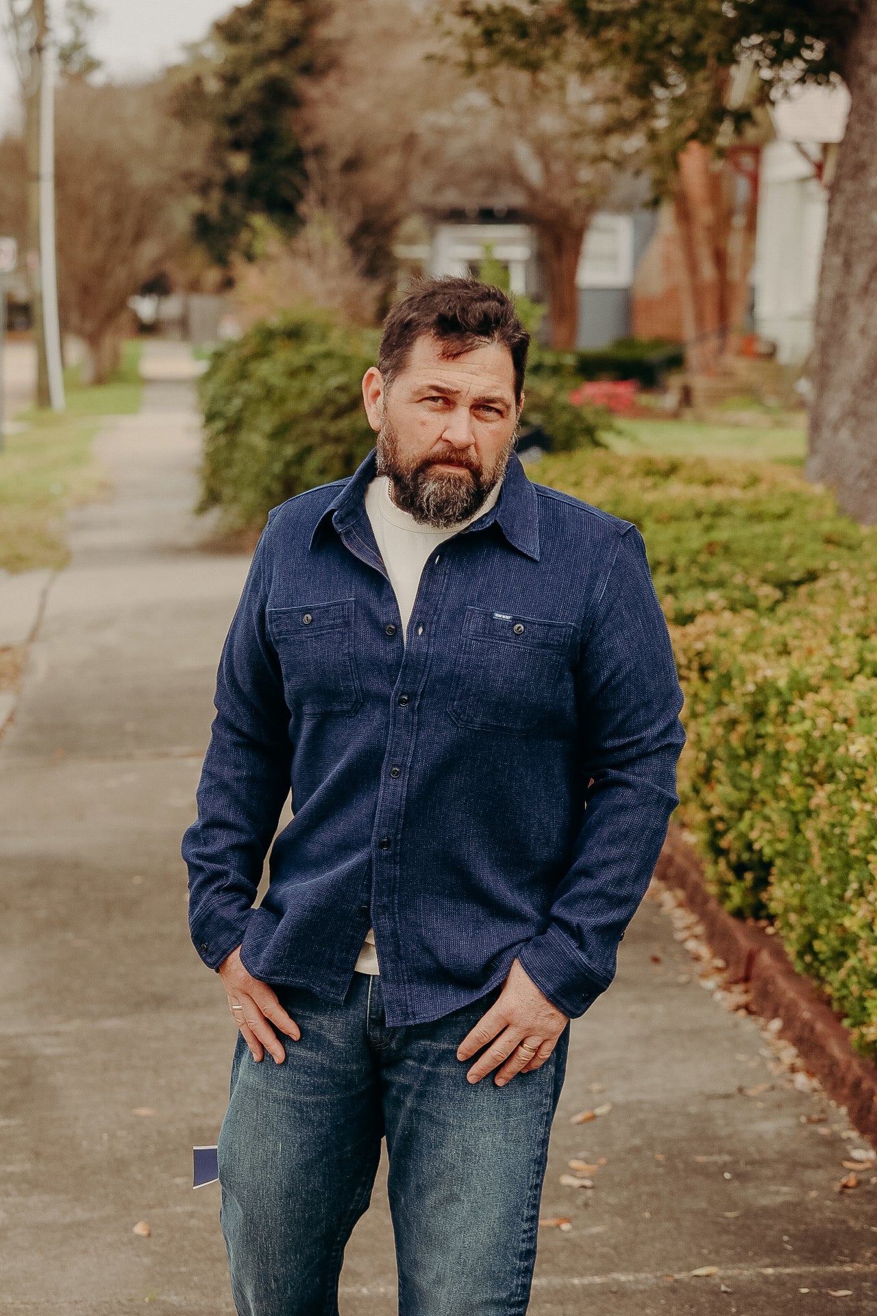 Bearded man wearing blue button-up shirt and jeans standing on sidewalk with green bushes