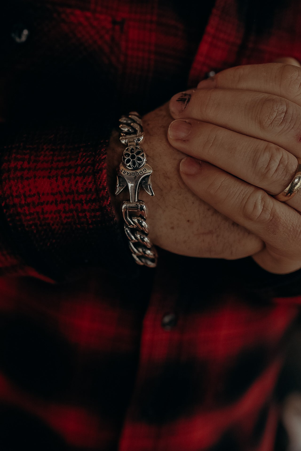 Close-up of hands wearing a silver chain bracelet engraved with 'GOOD ART' on a red and black plaid shirt