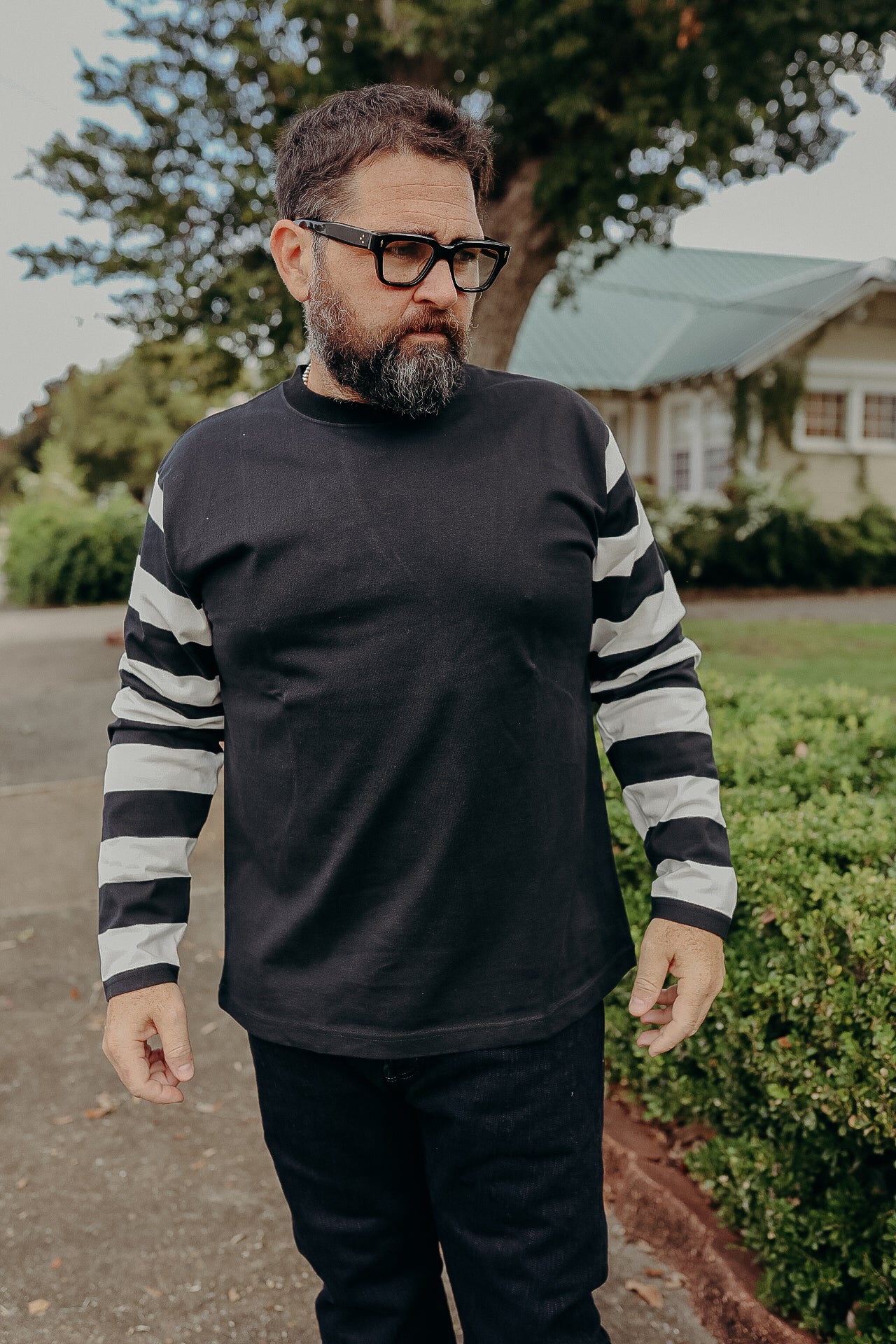 Man with beard and glasses wearing black shirt with black-and-white striped sleeves outdoors near a house