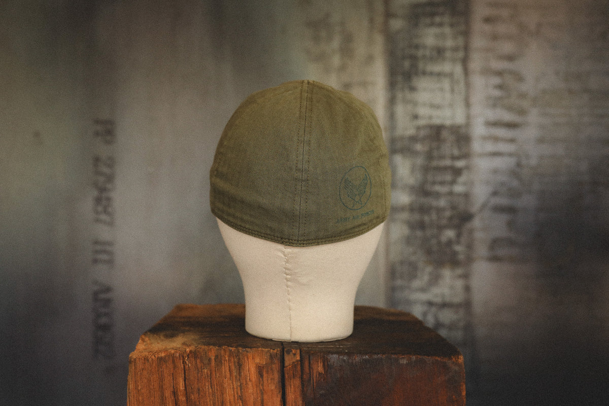 An olive green baseball cap displayed on a white mannequin head mounted on a rustic wooden block, set against a dark, textured gray background.