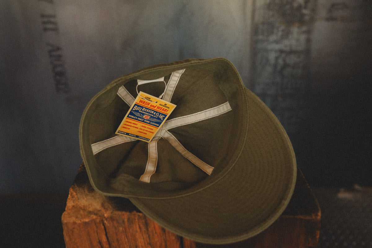 An olive green cotton flat-brim cap with white elastic cross-straps and a yellow and blue 'The Historic Hats' hangtag, resting on a rustic wooden stump against a dark, textured background.