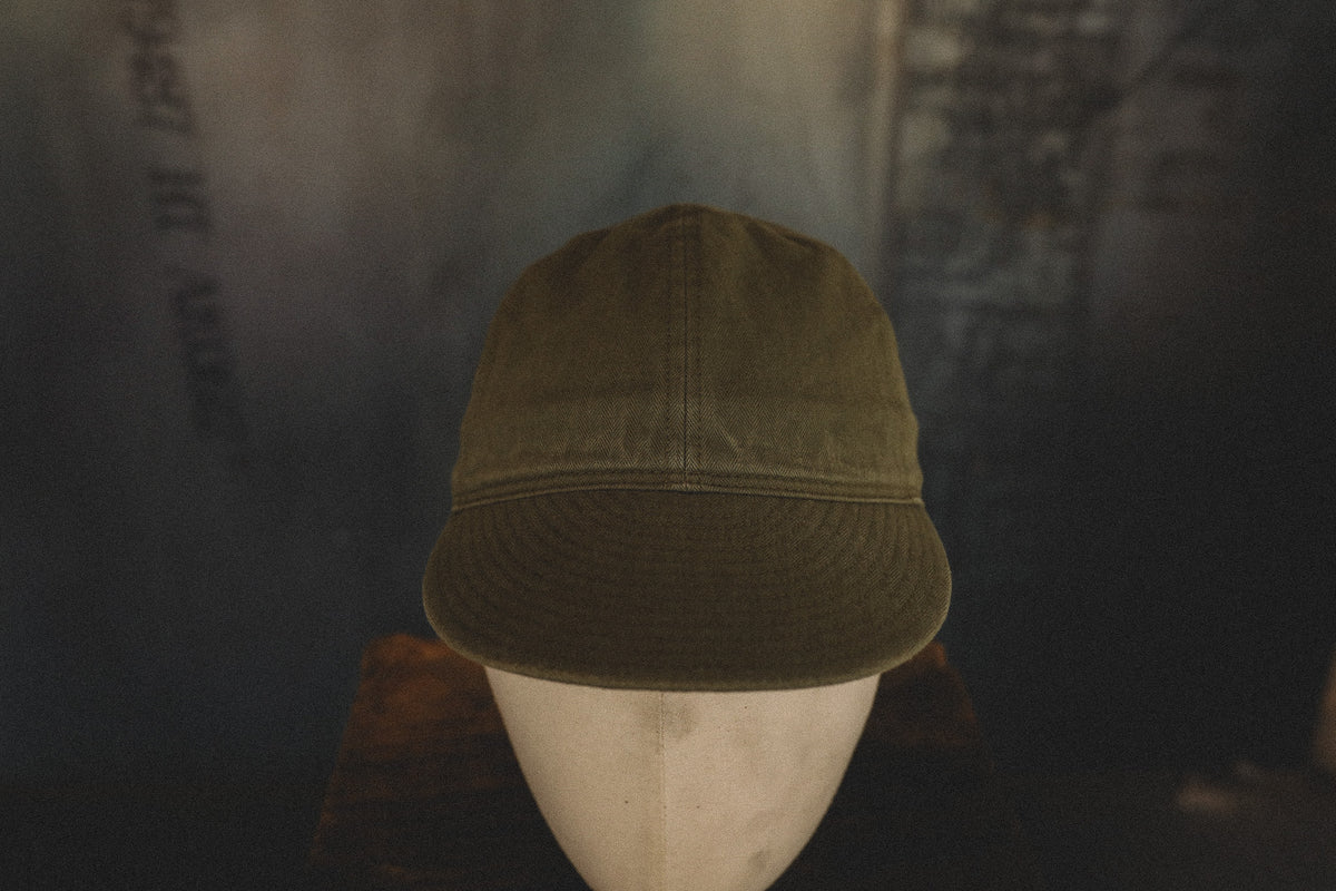 An olive green baseball cap displayed on a white mannequin head against a dark, moody background.