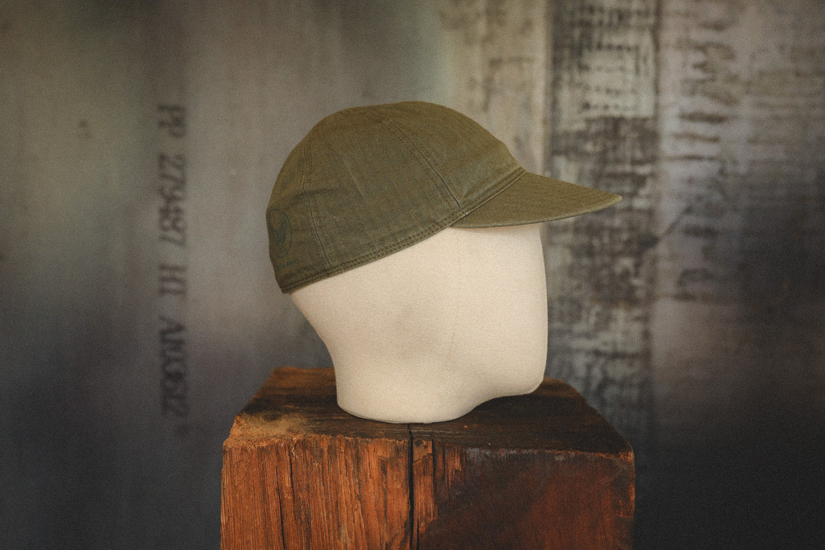 An olive green baseball cap displayed on a cream-colored mannequin head, which sits on a rustic wooden block against a textured, dark gray background.