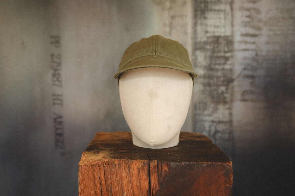 An olive green baseball cap displayed on a white fabric mannequin head, which sits on a rustic wooden block against a textured, dark gray background.