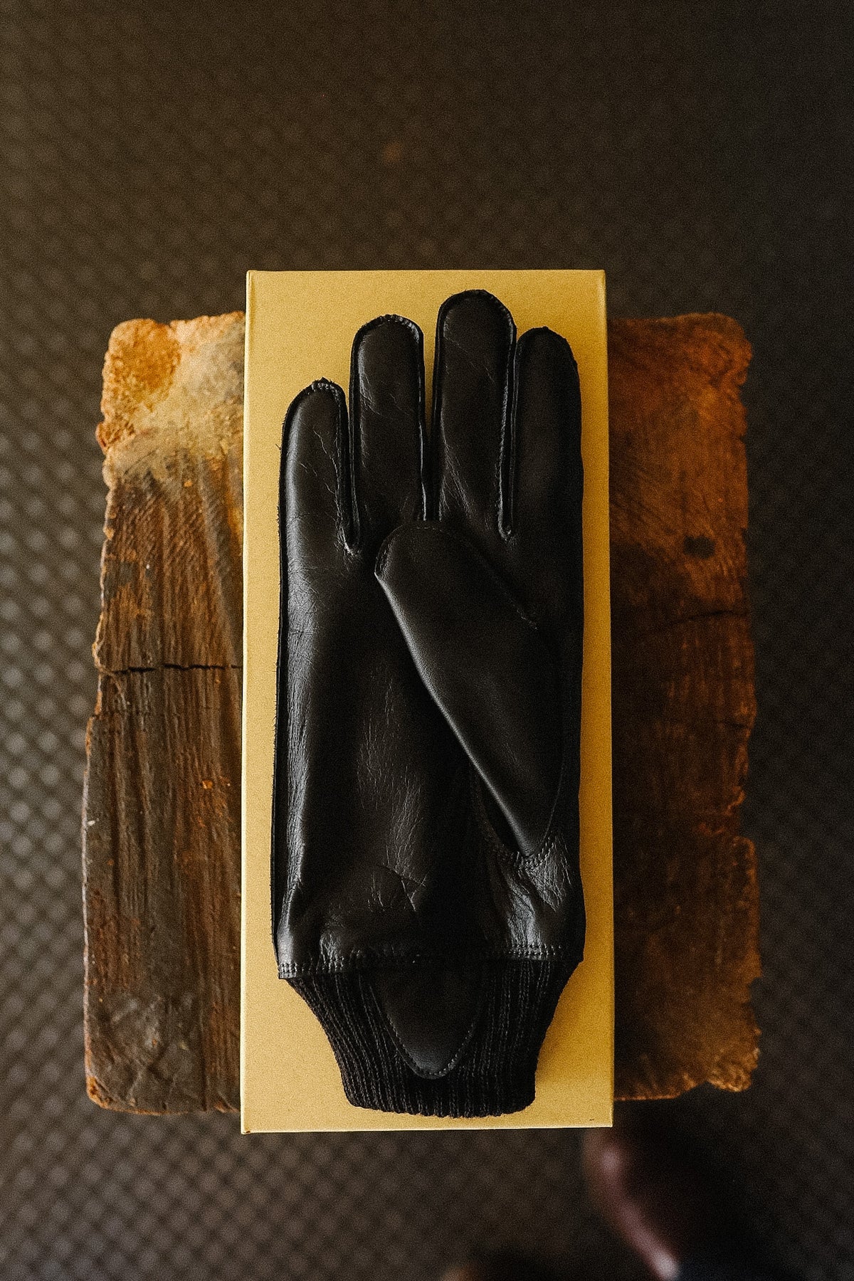 A single black leather glove laid flat on a rectangular tan box, with rustic wooden blocks behind it on a dark textured surface.