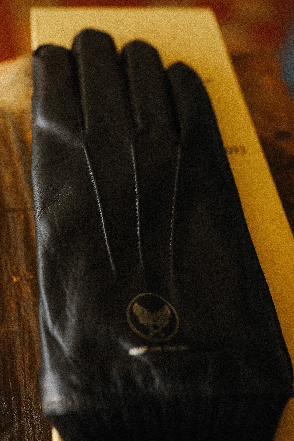 A black leather glove with a circular eagle logo near the cuff, resting on a yellow box over a wooden surface.