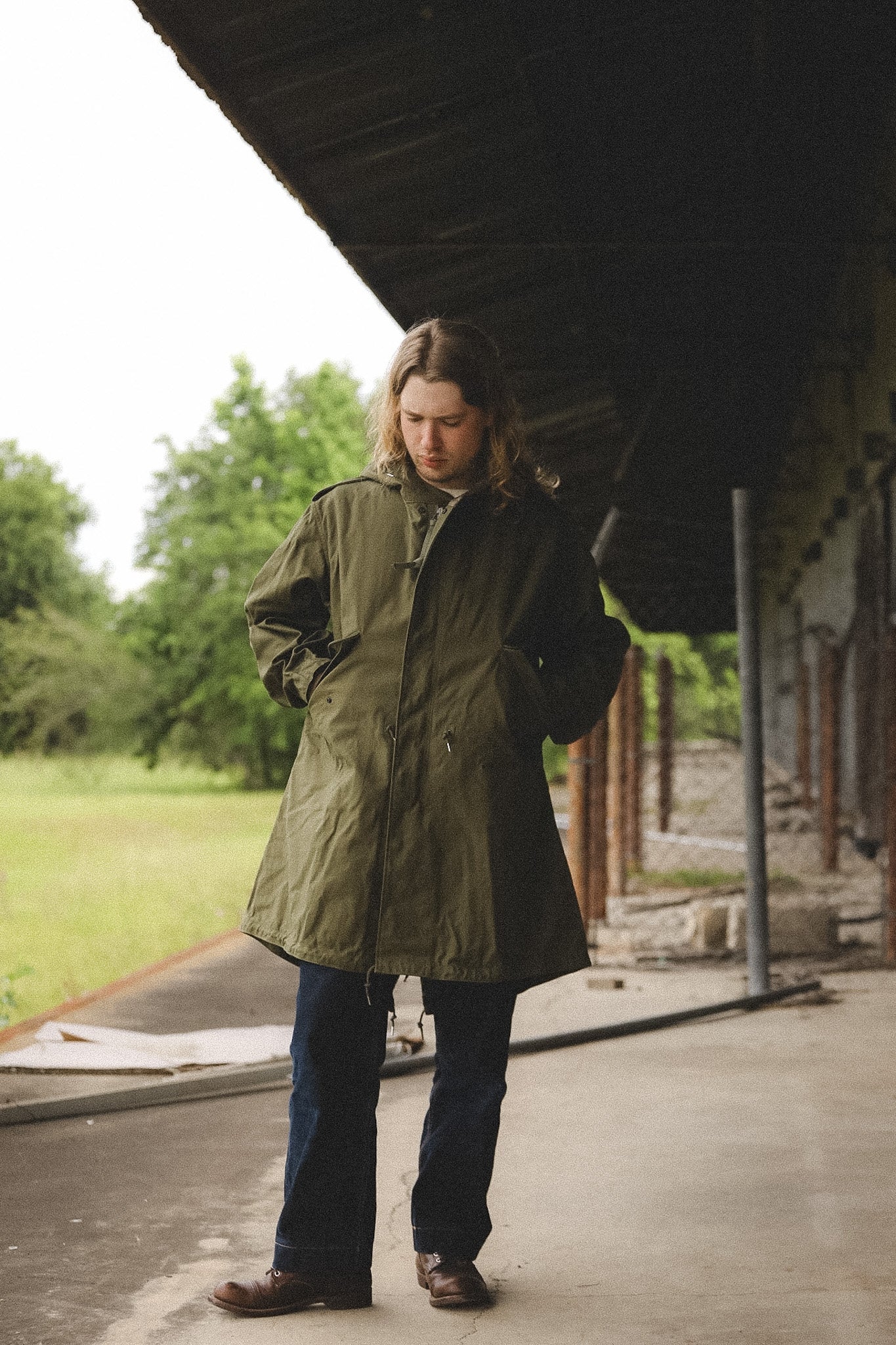 Person wearing a green coat standing under a large structure with trees in the background