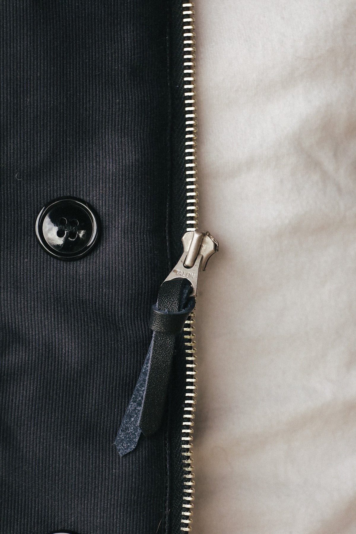 Close-up detail of a dark charcoal corduroy jacket with a black button, a gold-toned zipper, and a black leather zipper pull against a cream-colored textured fabric background.