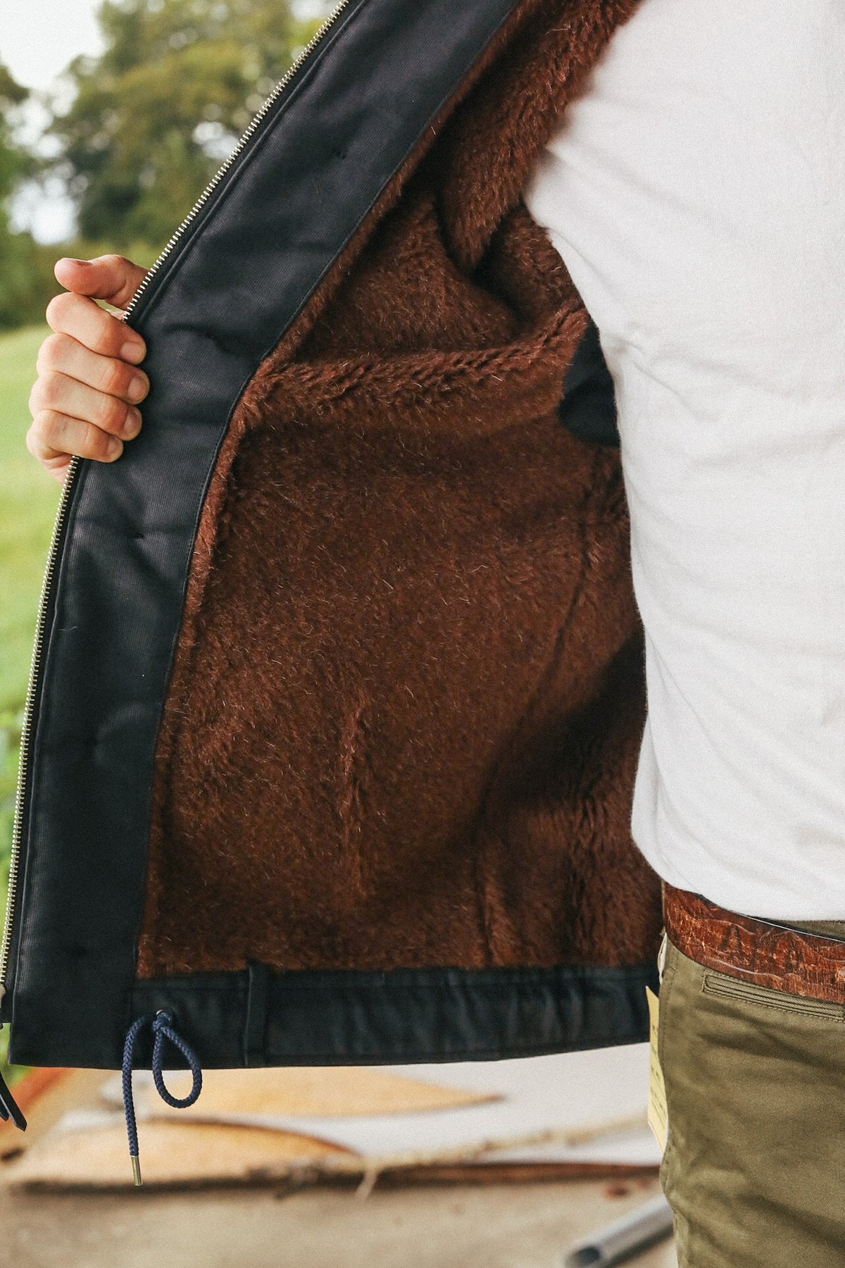 A close-up of a person wearing a black jacket with a rich brown sherpa lining, shown as they open the jacket to display the interior. The person is also wearing a white t-shirt and khaki pants.