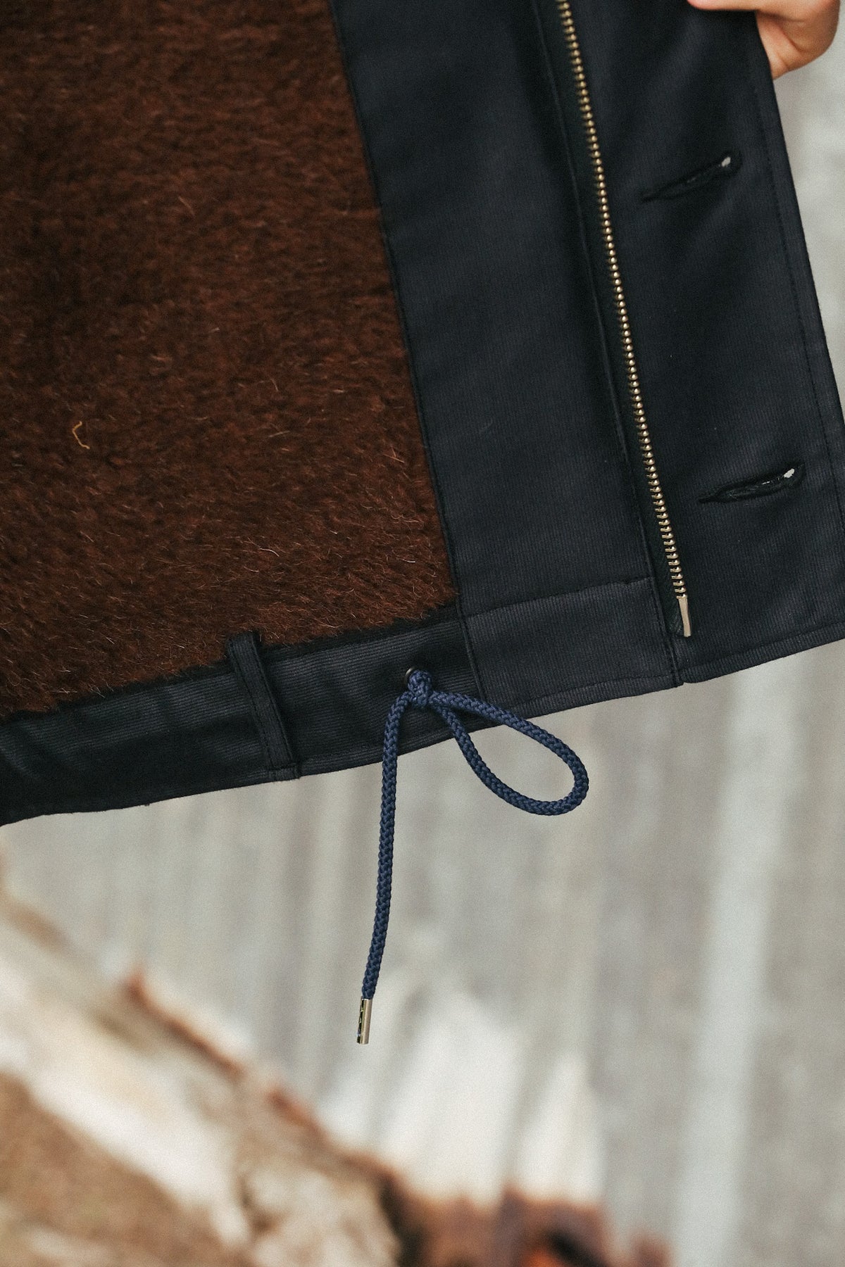 Close-up of a dark navy jacket or coat with a brown textured fleece or wool lining, a gold-tone zipper, and a navy drawcord, held by a hand against a blurred outdoor background.