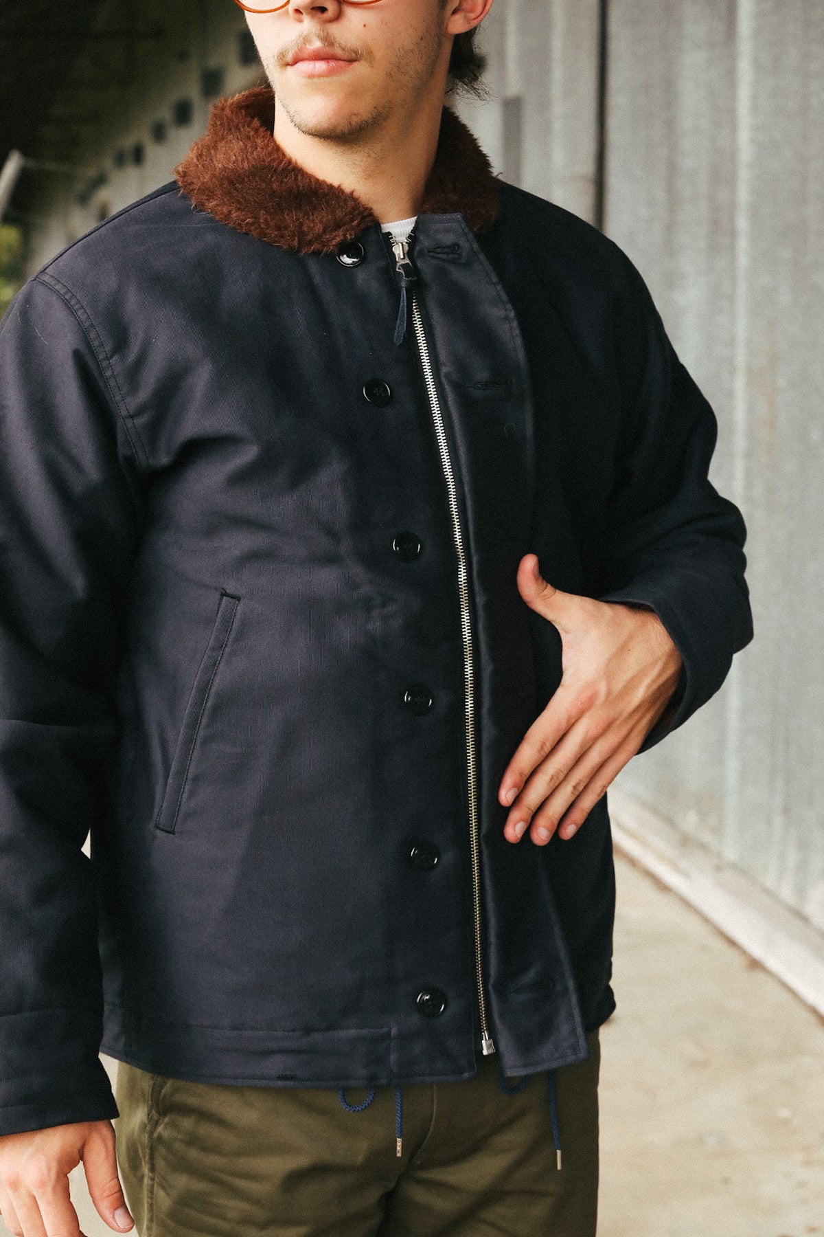 A man wearing a dark navy blue canvas jacket with a brown faux-fur collar, standing outdoors against a gray concrete wall.