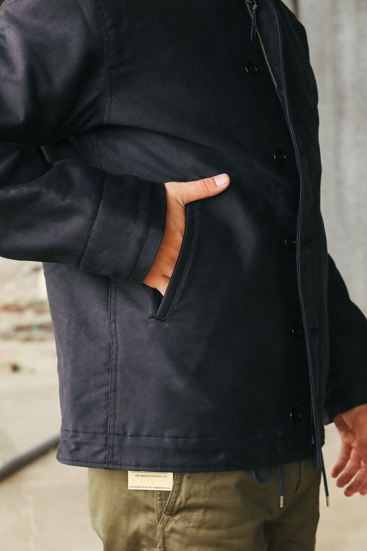 A close-up of a person wearing a dark navy utility jacket with a hand in the pocket, paired with olive green pants.