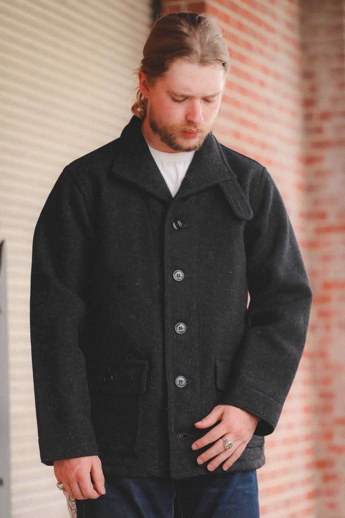 A person  wearing a dark charcoal gray wool-blend coat, standing in front of a red brick wall and a metal roll-up door.