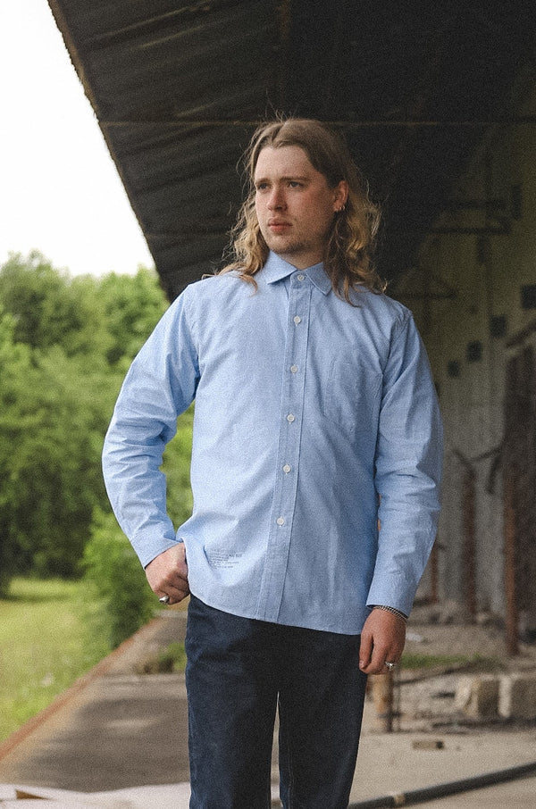 Man wearing a light blue shirt and dark pants standing on a platform with greenery in the background