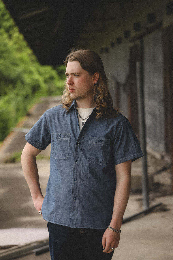 Man wearing a blue denim shirt standing outdoors with greenery in the background
