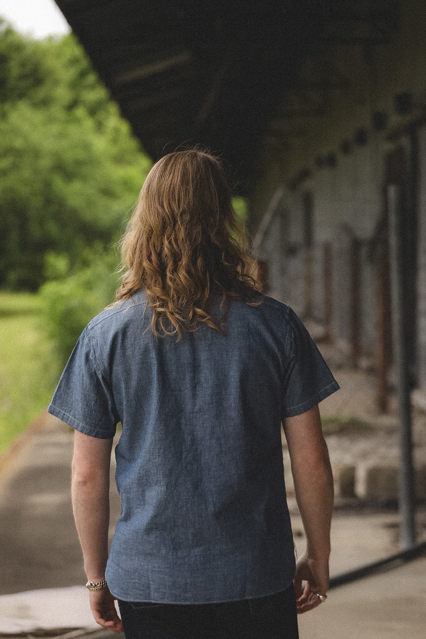 Person wearing a blue shirt standing on a path with greenery and a building in the background