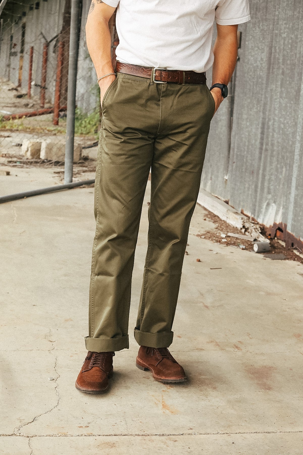 A person wearing olive green chino pants, a white t-shirt, a brown belt, and brown leather shoes, standing outdoors against a corrugated metal wall.